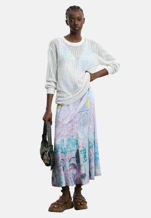 Kvinde stående iført en hvid gennemsigtig sweater, pastelblomstret midi-nederdel, brune platformsandaler og holder en mørk håndtaske med dekorative charms.