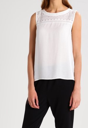 Mouwloze witte blouse met kantdetail over de bovenborst, gedragen met een zwarte broek, getoond van nek tot heupen.