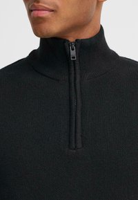 Svart strikket genser med høy krage, som har full glidelås. Stoffet har en teksturert overflate, som fremhever varme og komfort.