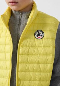 Gele opgevulde bodywarmer met doorgestikt ontwerp, voorzien van een ronde patch met tekst en een silhouet. Draagt over een grijze sweater met rits.