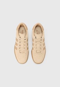 Beige mockasneakers med tre mörkare bruna ränder, rund tå, platta snören och en strukturerad sula. Insidan har en märkt innersula.