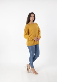 Mosterdgele oversized trui met geribbelde manchetten en onderzoom, gecombineerd met blauwe spijkerbroeken en heldere hoge hakken sandalen.