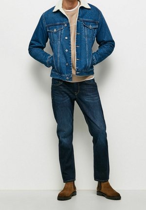 Mann trägt blaue Jeansjacke mit weißem Fleecekraus, beigefarbenes Sweatshirt, dunkelblaue Jeans und braune Wildlederstiefel, steht mit den Händen in den Taschen.