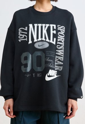 Osoba nosząca czarną bluzę Nike z białym i szarym tekstem zawierającym "1972," "Nike," "Sportswear" oraz "90," oraz wieloma logo Nike.