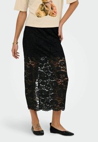 Falda de encaje negra con un patr�ón floral, que presenta una sección superior sólida y una parte inferior transparente. Combinada con zapatos planos negros.