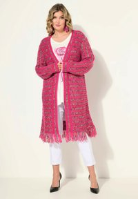 Szydełkowy kardigan w jaskrawym różowym kolorze z wielokolorowymi akcentami, z długim frędzlem u dołu, otwartym przodem i prążkowanymi mankietami, zestawiony z białymi spodniami.
