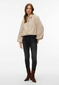 Veste en tricot beige avec fermeture éclair, manches surdimensionnées et col, associée à un jean skinny noir et des bottines marron.