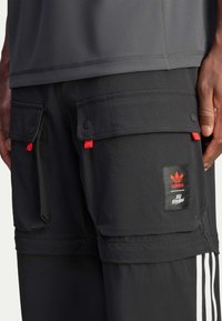 Black cargo shorts with red accents, featuring multiple pockets, snap fasteners, and an Adidas logo patch. Made from lightweight fabric.
