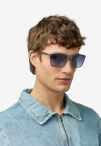 Pruunide ja siniste aktsentidega ristkülikukujulised päikeseprillid, tumenevate läätsedega ja matt viimistlusega, kantud hele sinise teksajaki seljas.