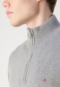Gros plan sur une personne portant un pull à fermeture éclair quart de longueur gris clair avec un petit logo rouge, blanc et bleu marine sur la poitrine.