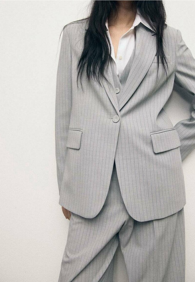 Personne portant un costume gris clair à fines rayures avec une veste croisée sur une chemise blanche à col, debout avec une main sur la hanche.