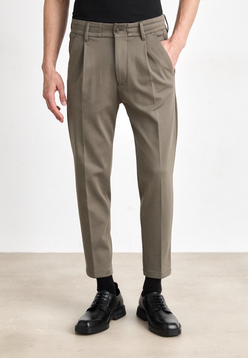 Pantaloni khaki con pieghe, chiusura frontale con bottone e tasche laterali, abbinati a scarpe e calzini neri, mostrati su uno sfondo neutro.