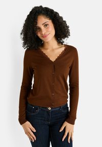 Jeune femme aux cheveux bouclés portant un cardigan marron à boutons et un jean bleu foncé, se tenant devant un fond clair et uni.