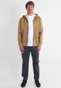 Un sweat à capuche marron zippé avec un intérieur gris clair, un cordon de serrage et des poches latérales, porté par-dessus un t-shirt blanc et associé à un pantalon gris foncé.