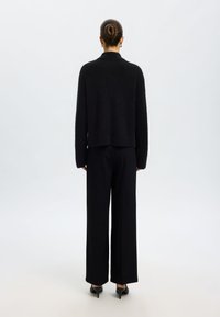 Crni pahuljasti džemper dugih rukava s okruglim ovratnikom, uparen s crnim hlačama širokih nogavica visokog struka. Glatka tekstura, minimalne detalje.