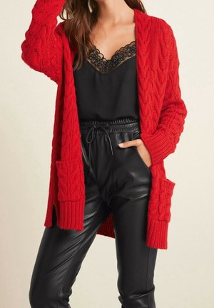 Femme portant un haut noir bordé de dentelle, un pantalon en cuir noir taille haute et un long cardigan rouge en maille torsadée avec poches.