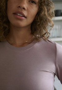 Žena s kovrčavom kosom i svijetlosmešnim tenom nosi zlatni lančić i dugi mauve majicu s dugim rukavima u zatvorenom prostoru.