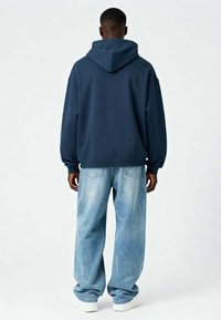 Marineblauwe hoodie met een ruim ontwerp, voorzien van een trekkoordkap, ribbels aan de mouwen en aan de achterkant. Gecombineerd met loszittende, lichtgekleurde spijkerbroeken.