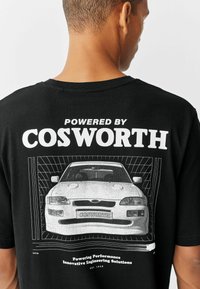 Camiseta de algodón negra con un gráfico de un coche blanco en la parte trasera, con el texto "Powered by COSWORTH" y texto adicional en fuente blanca.