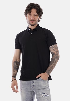 Uomo con capelli ricci che indossa una polo nera e jeans azzurri strappati, con tatuaggi dettagliati su entrambe le braccia, mano in tasca.