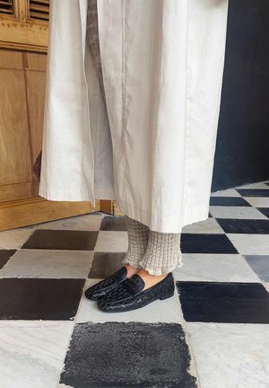 Personne portant un pantalon large de couleur crème, des leggings en maille beige et des mocassins en cuir tressé noir, debout sur un sol en carreaux noirs et blancs.