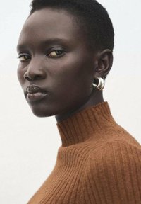 Suéter marrón de cuello alto con costillas y tejido texturizado. El modelo tiene piel oscura, cabello corto y pendientes circulares plateados, mirando directamente a la cámara.