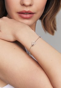 Srebrna bransoletka z wzorem serca i małymi, przezroczystymi akcentami, przymocowana do delikatnego łańcuszka. Ramię modelki jest częściowo widoczne.