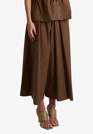 Donna che indossa pantaloni larghi marroni e sandali beige con tacco alto e spesse fasce, in piedi su sfondo bianco.