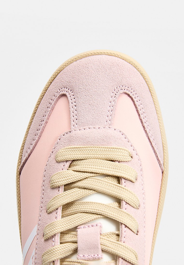 Zapatilla deportiva rosa claro con materiales de ante y sintéticos, con cordones beige y suela de goma texturizada. Detalle de rayas laterales blancas.