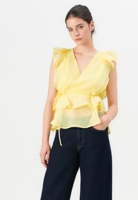 Top amarillo con volantes en las mangas, hecho de una tela ligera. Presenta un cinturón en la cintura y un diseño en capas, combinado con jeans de pierna ancha oscuros.