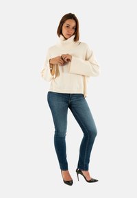 Mujer vestida con un suéter de cuello alto oversize color crema, jeans ajustados azules, tacones altos negros, de pie con las manos entrelazadas y una expresión neutral.