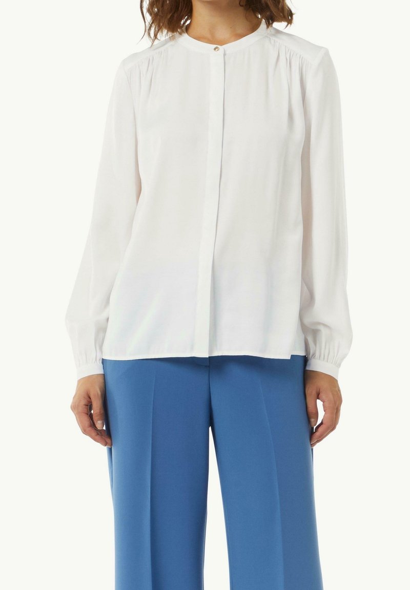 Femme portant un chemisier blanc à manches longues avec des poignets boutonnés et un pantalon bleu taille haute à jambes larges, debout devant un fond uni.