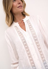 Vit blus med volangkrage, som har delikata knipplade insatser, knappstängning framtill och strukturerade ränder på tyget. Långa, något puffiga ärmar.