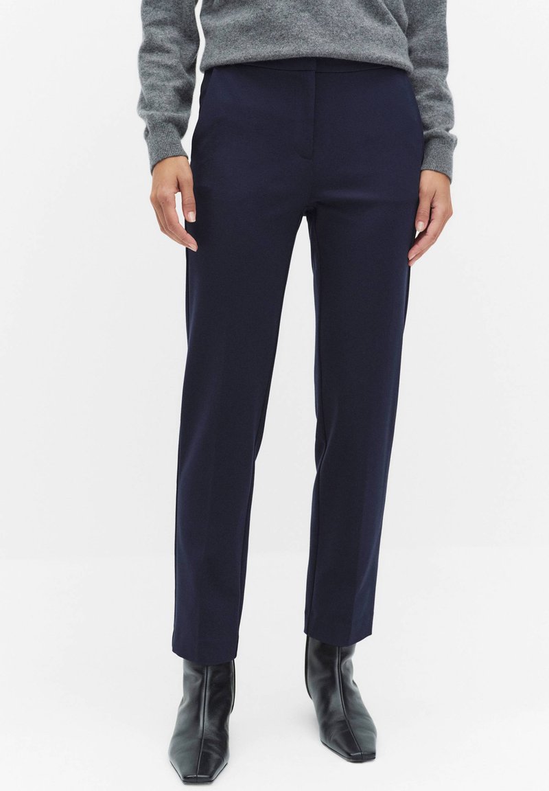 Personne portant un pantalon bleu marine sur mesure, un pull gris et des bottines noires à bout pointu, debout devant un fond blanc uni.