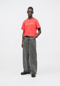 T-shirt vermelha de manga curta com o logótipo "marimekko" em bege, combinada com jeans cinzentos escuros oversize. Sapatos pretos completam o conjunto.