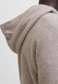 Sudadera beige con capucha y una textura suave, presenta un corte holgado y detalles de costura visibles a lo largo de las costuras y bordes.