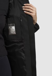 Schwarze, isolierte Jacke mit glatter Außenseite, Reißverschlusstasche und einem Stoffetikett mit Pflegehinweisen. Weiche Innenauskleidung sichtbar.