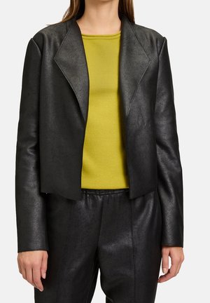 Kobieta ubrana w czarną skórzaną marynarkę i dopasowane spodnie, na żółto-zielonej koszulce z okrągłym dekoltem, stojąca na białym tle.