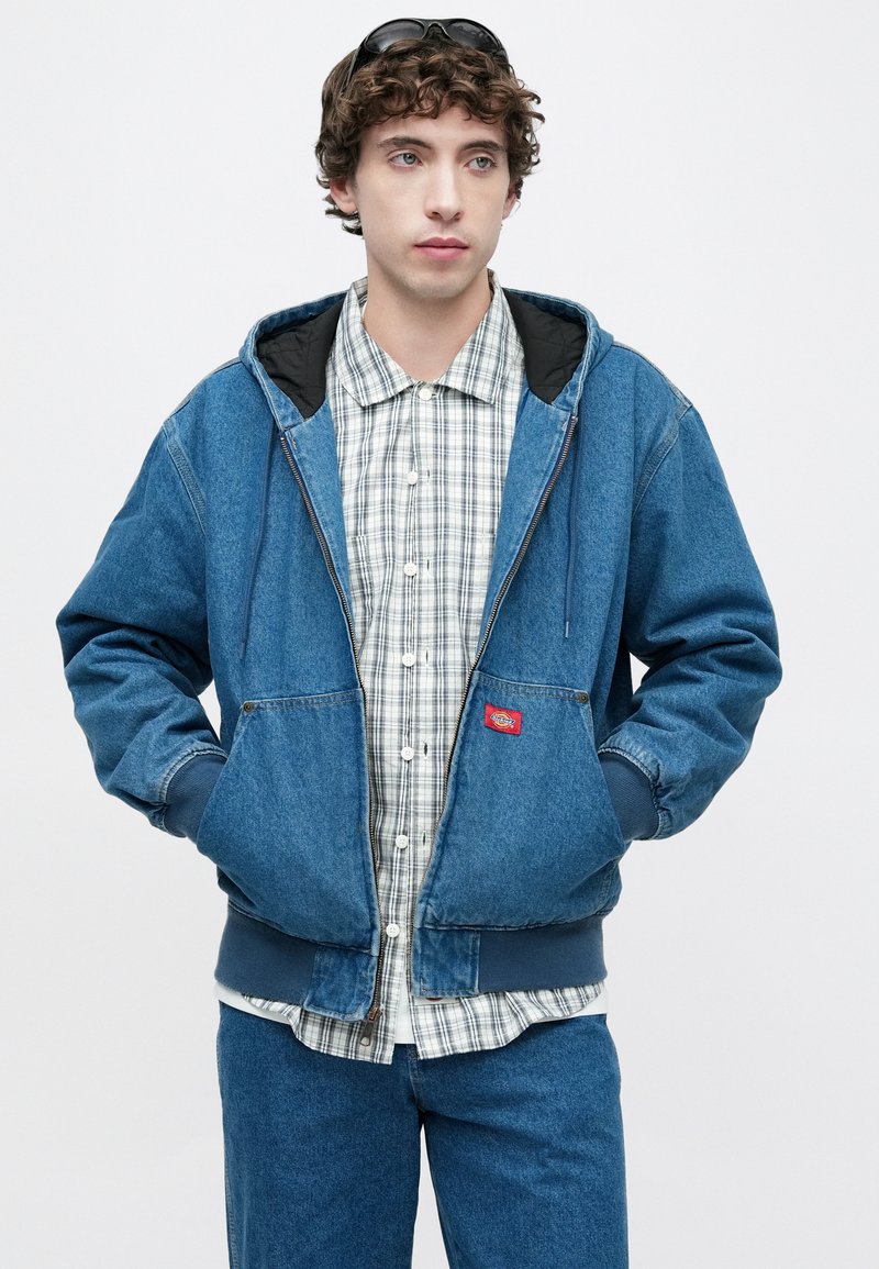 Blaue Jeans-Kapuzensjacke mit schwarzer Innenseite, ausgestattet mit einem Frontreißverschluss, zwei Taschen und einem Logo-Patch, getragen über einem karierten Hemd.