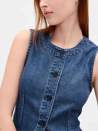 Woman wearing sleeveless blue denim button-up top, looking slightly to the side with straight red hair and small hoop earring.