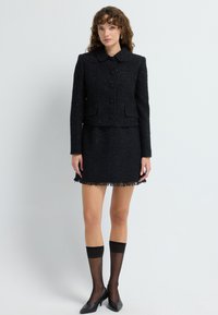 Sort tweedjakke med krave og fem knaplukninger, parret med en matchende nederdel med frynser i bunden. Sorte højhælede sko synlige.