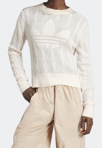 Jersey de punto en color blanco roto con diseño de trenzas, con un logo central. Puños y dobladillo acanalados, acompañado de pantalones ligeros en color beige.