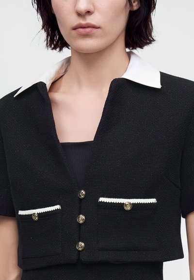 Femme portant une veste noire texturée à manches courtes avec un col blanc et des boutons dorés, par-dessus un haut côtelé noir.