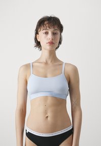 Lyseblå stretch-bralette med tynne justerbare stropper og en band med logo. Kombinert med svarte truser med en hvit logoband.