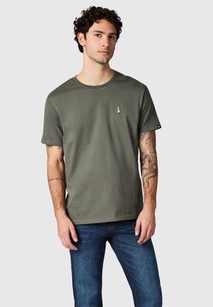 Giovane uomo con capelli ricci che indossa una maglietta verde oliva a maniche corte con un piccolo disegno di bottiglia sul petto e jeans blu, in piedi su uno sfondo grigio.