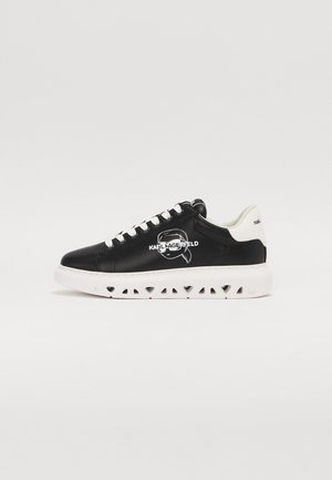 Black leather sneakers with white soles, featuring a logo on the side and a perforated sole design. Lace-up closure with white laces.