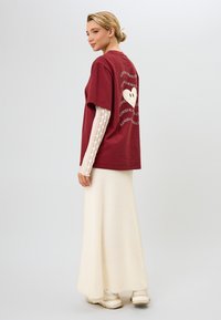 Burgundska oversize majica s belim grafičnim dizajnom na hrbtu. Slojevito oblečena z belo čipkastimi dolgimi rokavi in v kombinaciji s kremnim dolgim krilom.