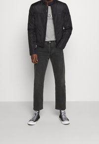 Giubbotto bomber nero con tasche con zip, t-shirt grigia a girocollo, jeans grigi e sneakers alte nere con suola bianca.