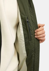 Giacca verde oliva con texture trapuntata, dotata di una tasca interna con zip nera e fodera color crema, tenuta aperta da una mano.