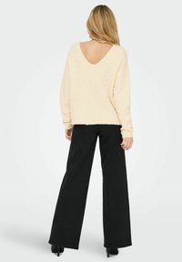 Cremfarbener, flauschiger Pullover mit tiefem V-Ausschnitt auf der Rückseite, kombiniert mit weit geschnittenen schwarzen Hosen und hohen Schuhen. Weiche Textur und lässige Passform.